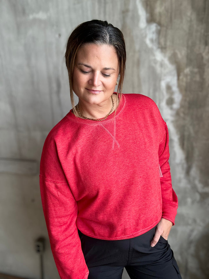 A woman wearing a heathered red long-sleeve crewneck sweatshirt and black pants, standing against a textured concrete wall.