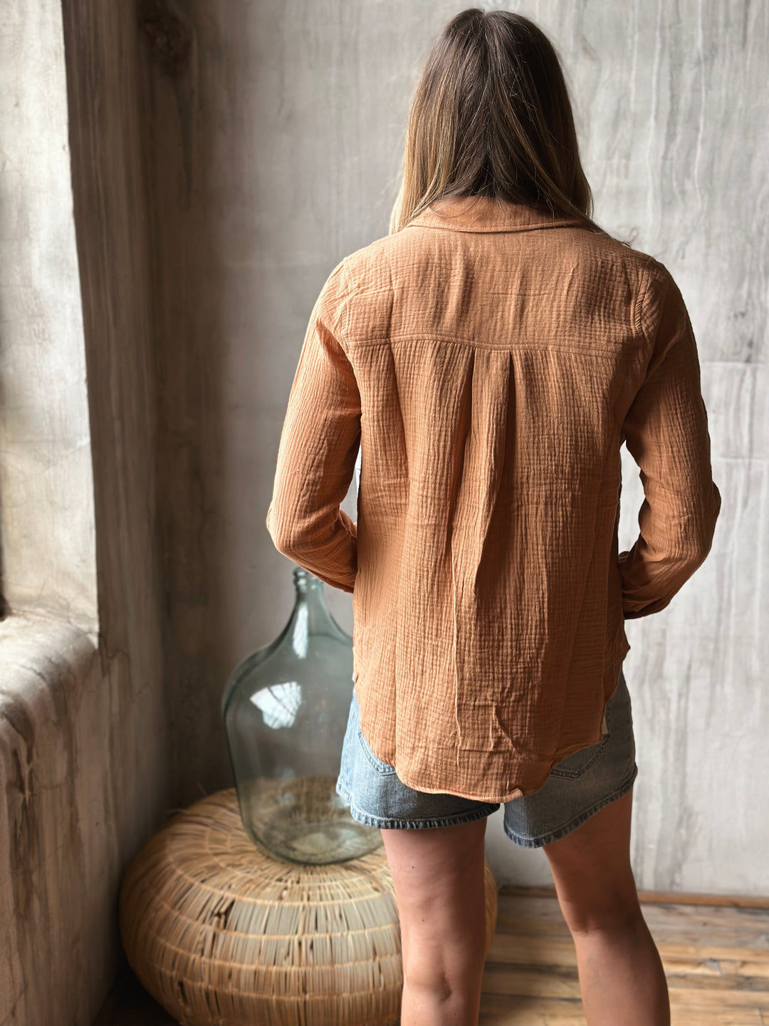 Woman wearing a burnt orange blouse in a room with a textured wall and large vase.