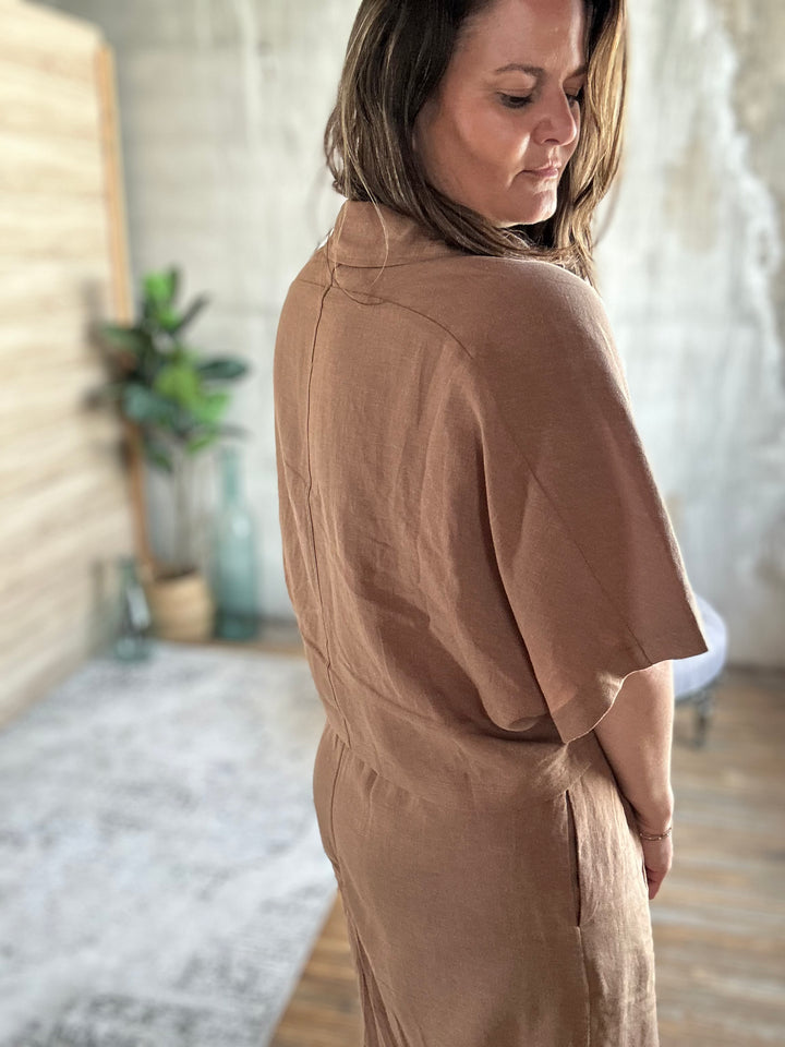 A woman wearing a brown outfit in a room with a plant and light-colored walls.