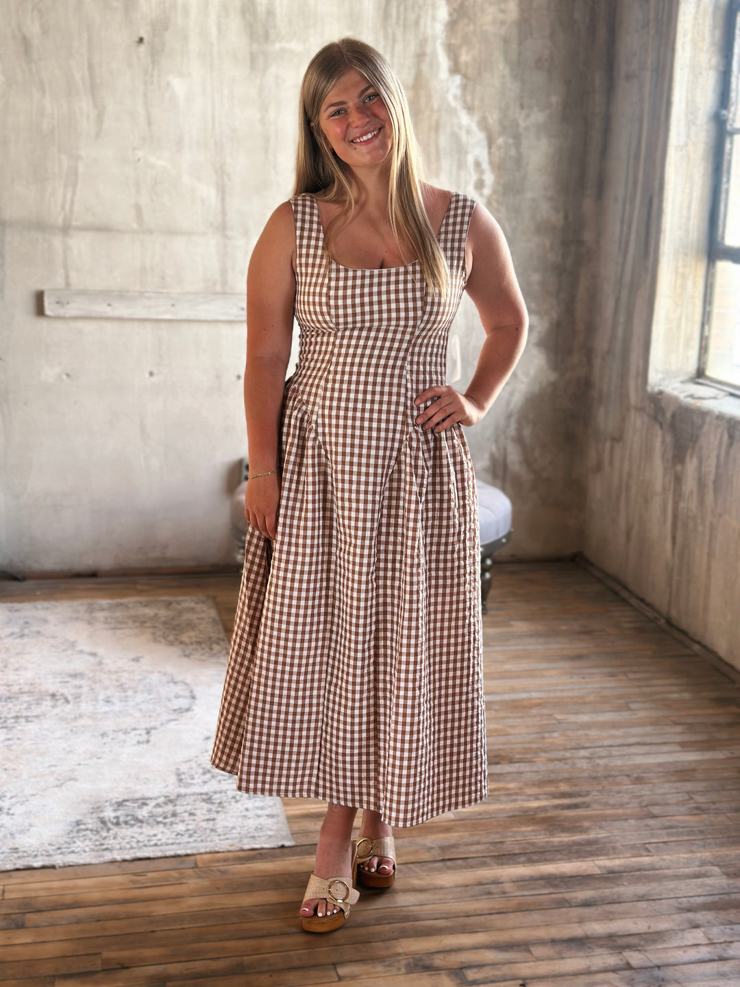 Woman wearing a brown and white plaid  dress standing in a room with a wooden floor and concrete wall.