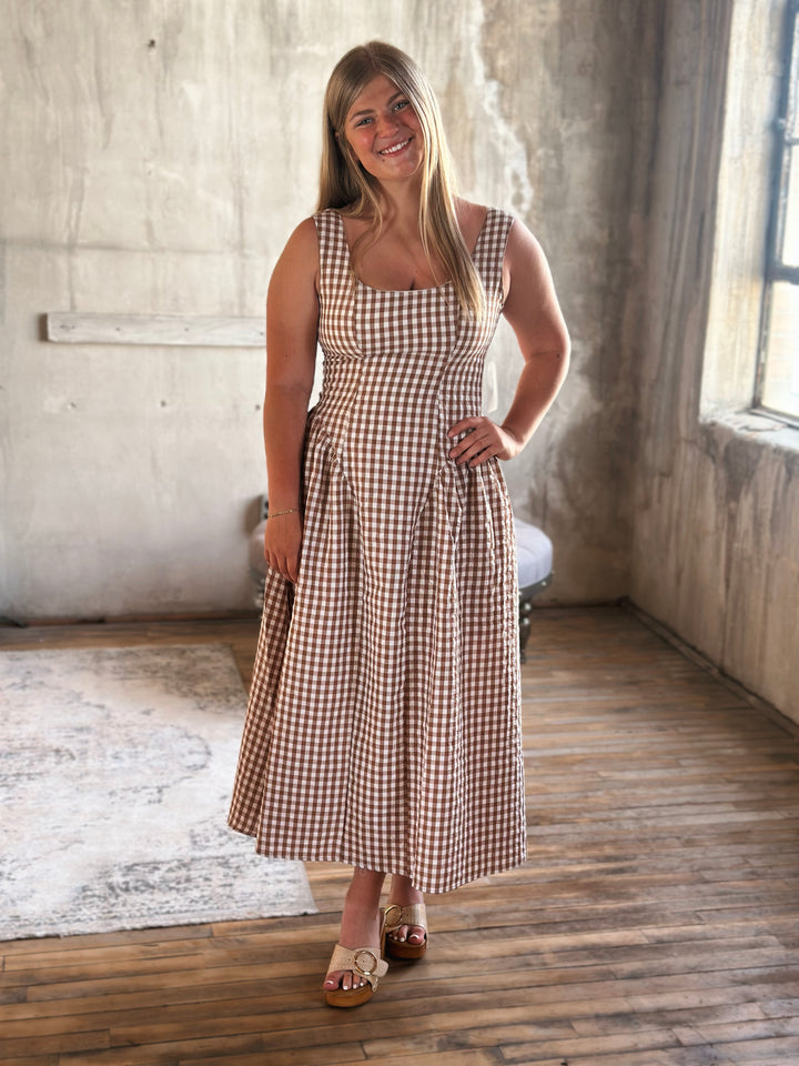 Woman wearing a brown and white plaid  dress standing in a room with a wooden floor and concrete wall.