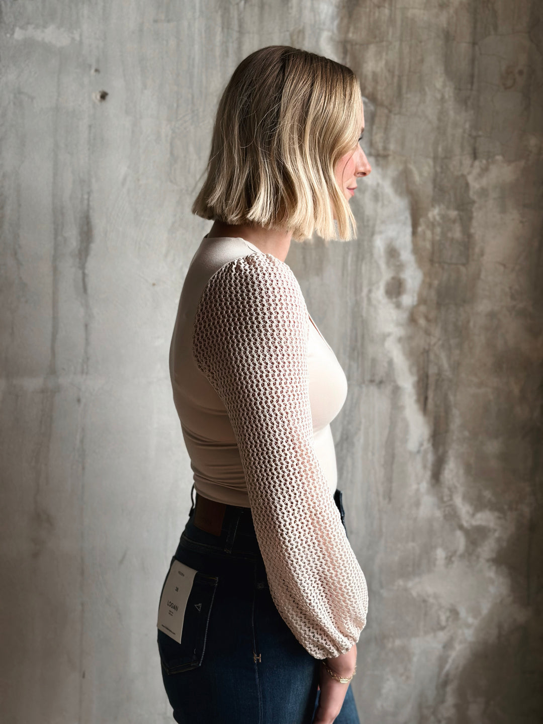 Woman wearing a beige knitted sweater against a textured wall.
