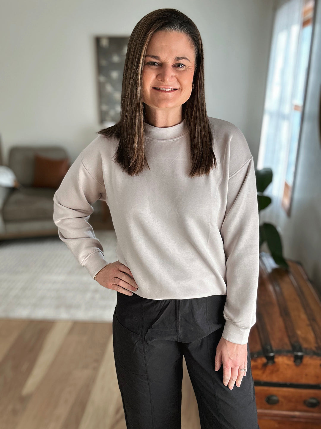 Woman wearing a light gray sweater and black pants in a room with a couch and window.