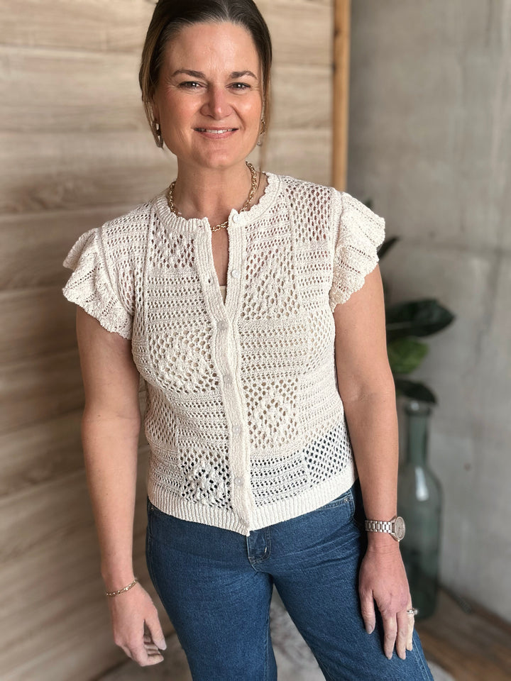 Woman wearing a white crochet top and blue jeans standing against a neutral wall.