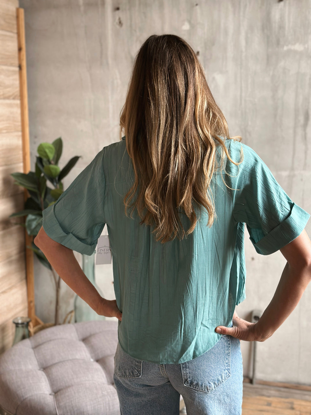 Woman wearing a teal blouse and jeans standing in a room with a plant and gray wall.