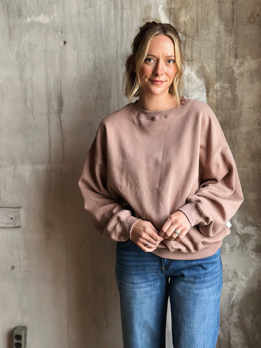 Woman wearing a pink sweater and blue jeans against a textured wall.