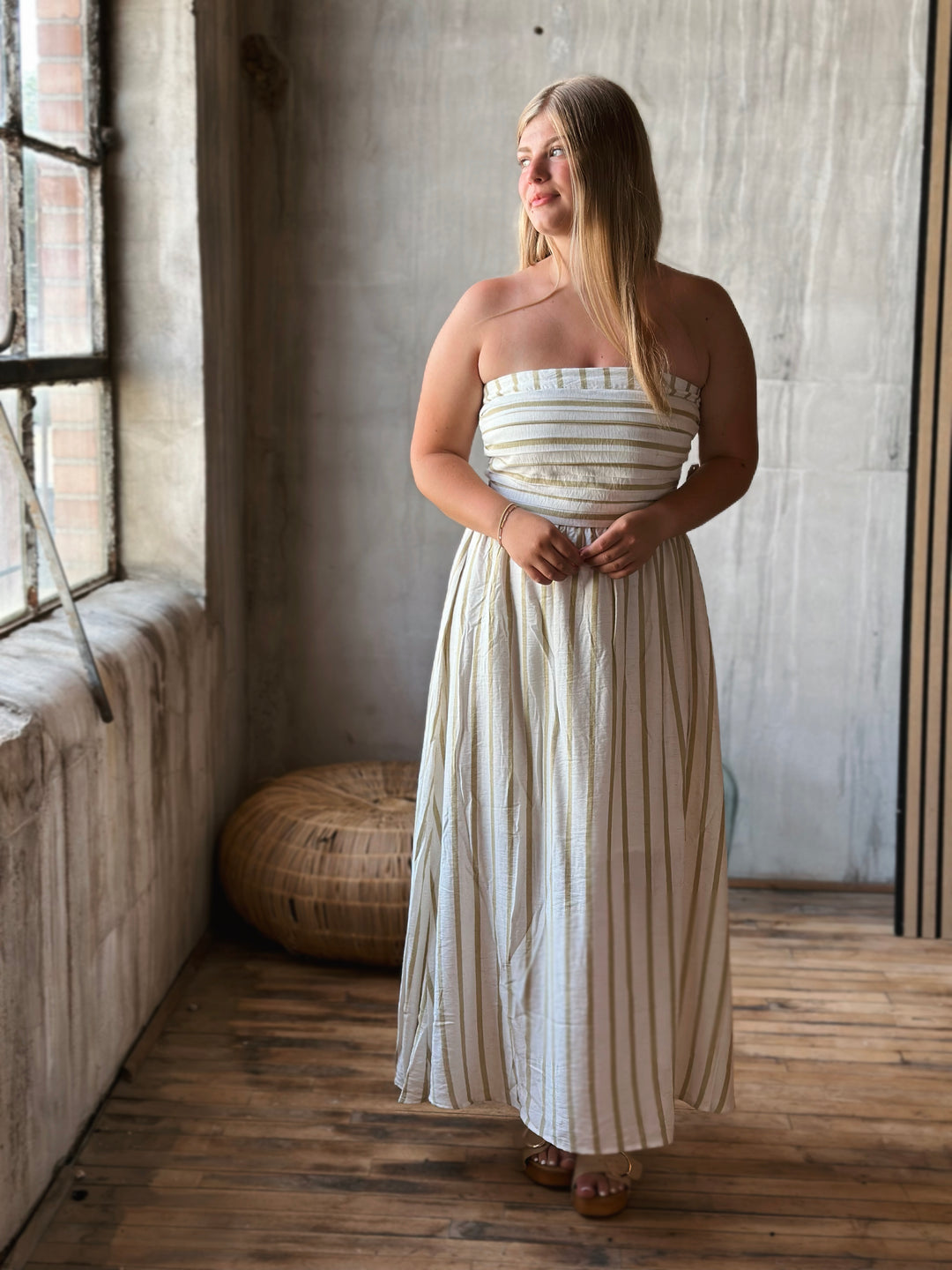 A woman wearing a long white strapless dress with beige stripes standing in a room with a wooden floor and concrete wall.