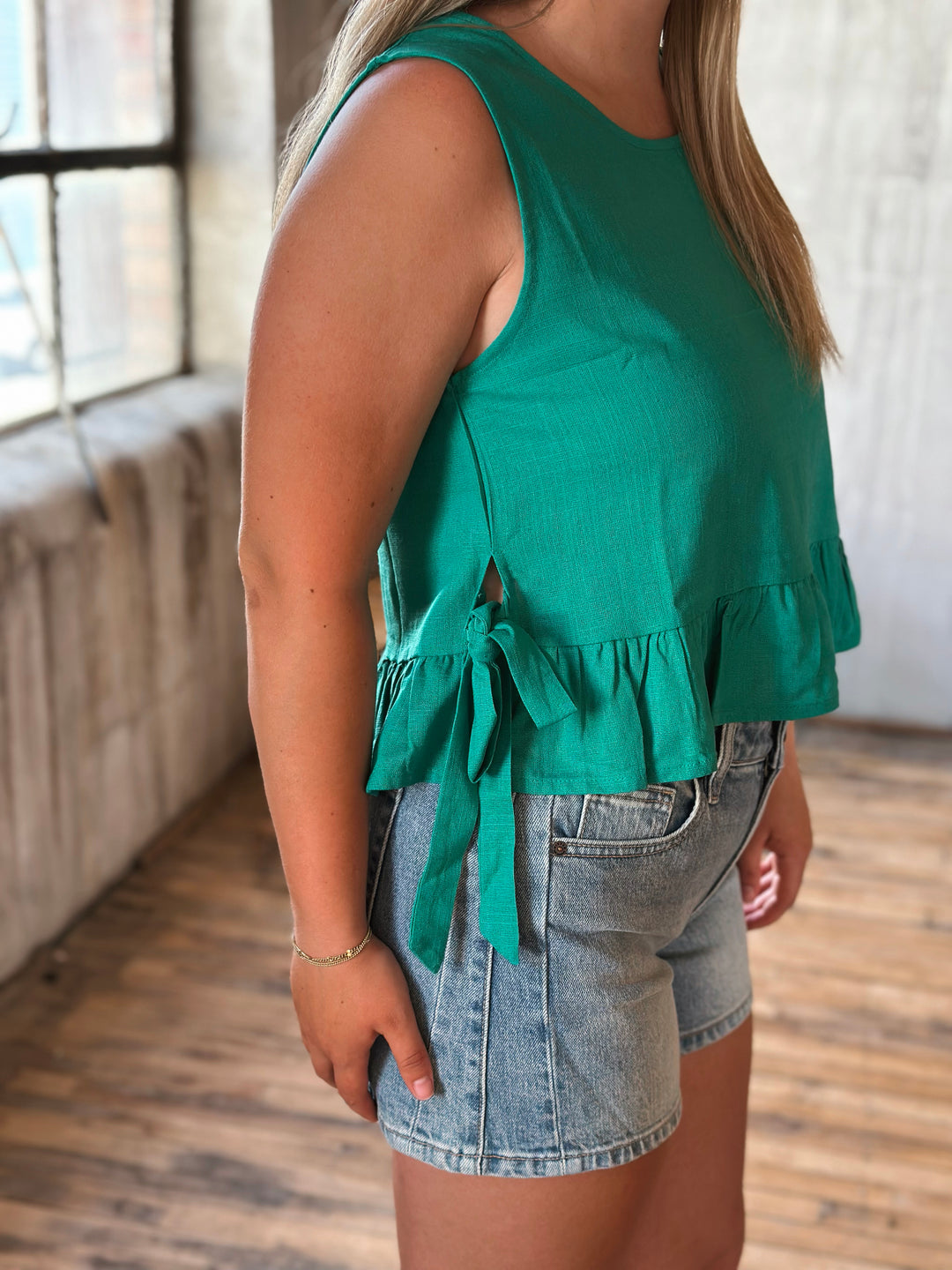 Person wearing a green sleeveless top with ruffle details and denim shorts indoors.