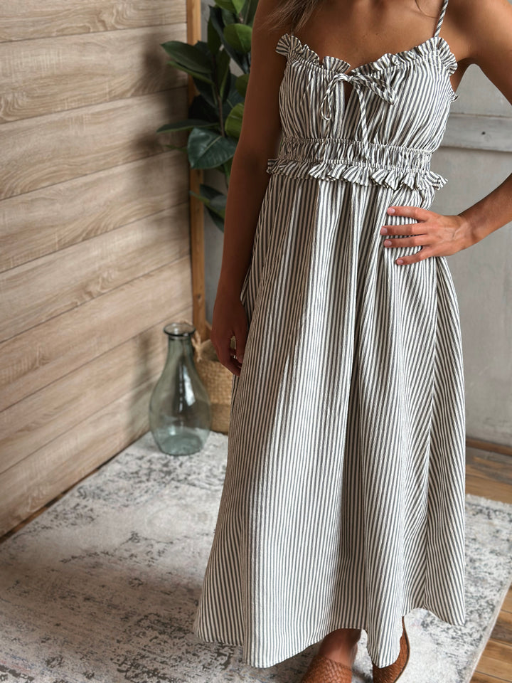 Woman wearing a white and gray striped dress standing indoors with a wooden wall and plant in the background.