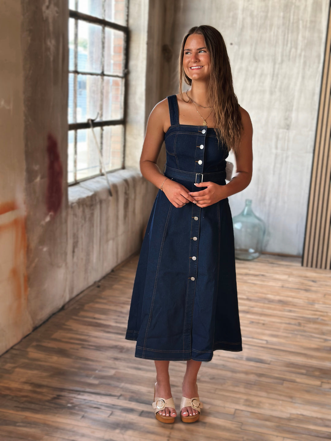 Woman wearing a blue denim dress with silver buttons in a room with wooden floor and textured walls.