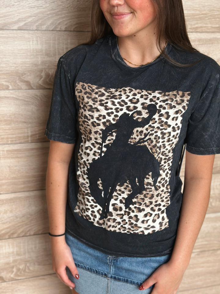 Person wearing a black t-shirt with a leopard print and silhouette of someone on a bucking horse against a wooden background.