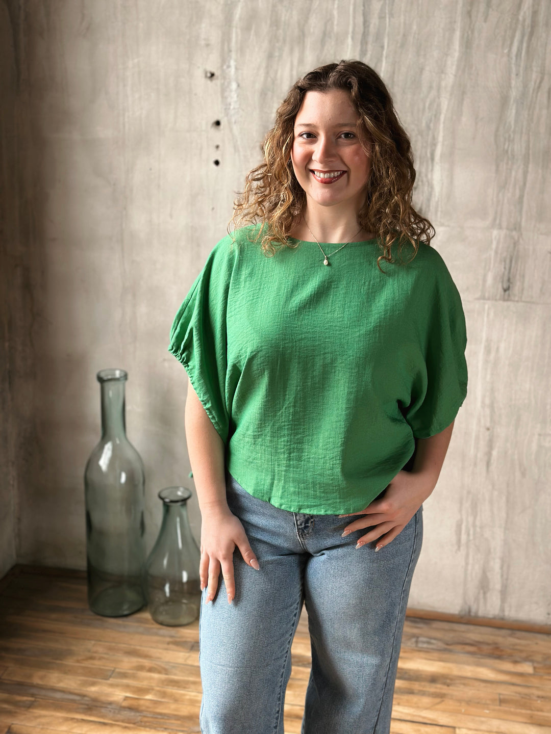 Woman wearing a green top and blue jeans standing in a room with wooden floor and textured wall.