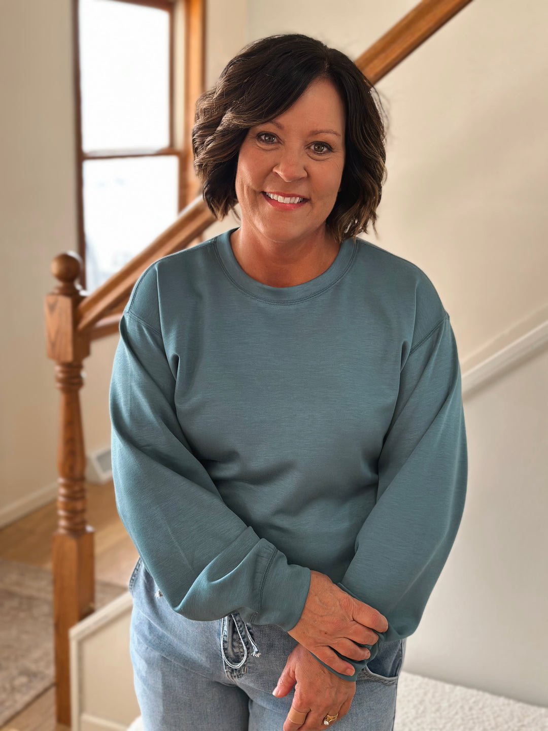 Woman wearing a blue sweatshirt standing in a home setting with a staircase.