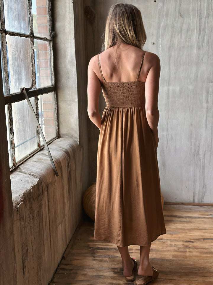 Woman in a brown dress standing by a window in a room with wooden floor and concrete wall.