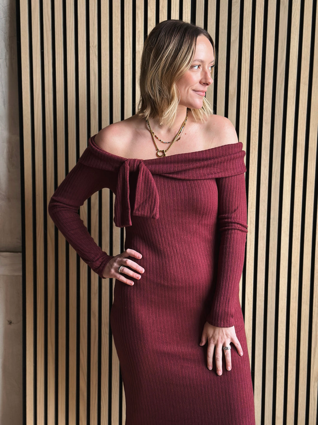 Woman wearing a burgundy off-shoulder dress against a wooden paneled wall.