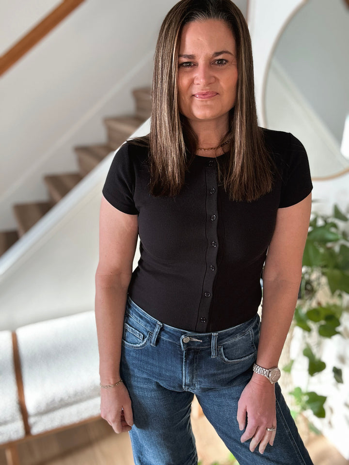 Woman wearing a black shirt and blue jeans standing in a home setting with stairs and plants.