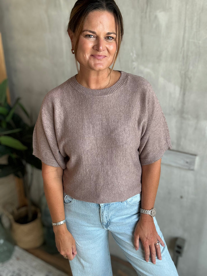 A woman wearing a short-sleeve knit sweater in a dusty mauve-brown color, paired with light-wash blue jeans.