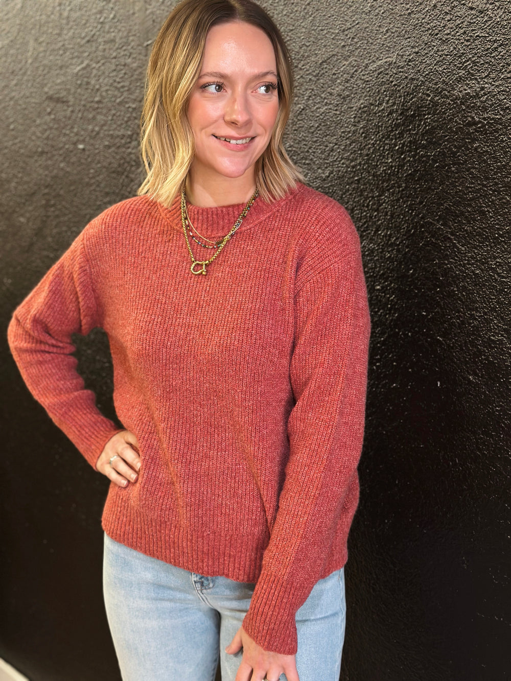 Woman wearing a rust-colored sweater against a dark textured wall