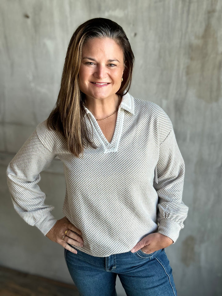 A woman with long brown hair smiling, wearing a light beige and white striped long-sleeve collared shirt with blue jeans, standing against a textured concrete wall.