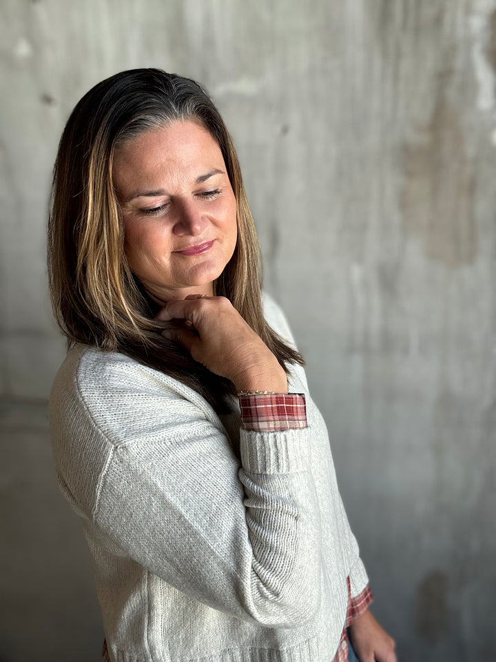 A woman with shoulder-length brown hair with blonde highlights, wearing a light gray knit sweater over a red plaid shirt, posing against a textured concrete wall.