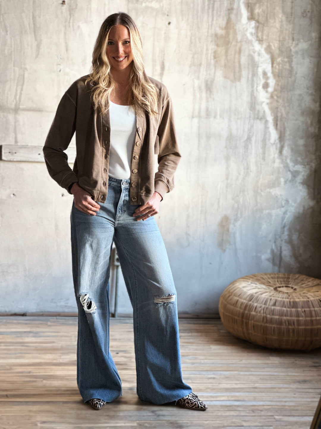 Woman wearing a brown jacket, white shirt, and blue jeans standing in a room with a textured wall and wooden floor.