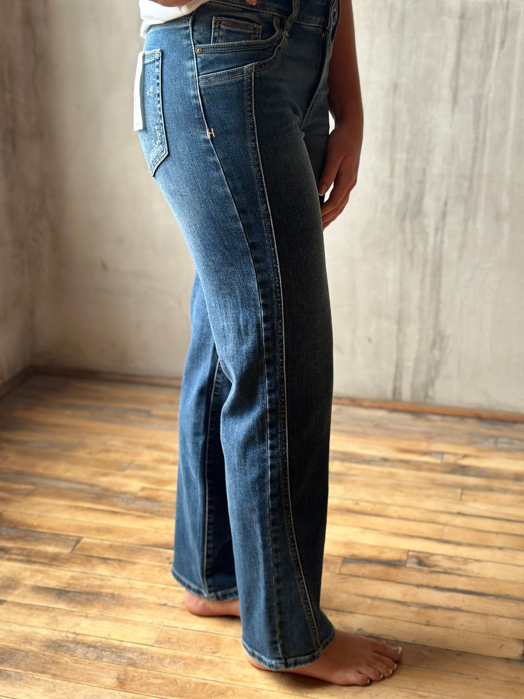 A person wearing dark blue denim jeans with a slight flare, standing barefoot on a wooden floor against a textured concrete wall.