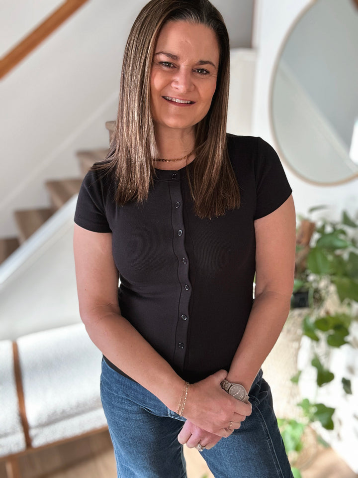 Woman wearing a black shirt and blue jeans standing indoors with a staircase in the background.