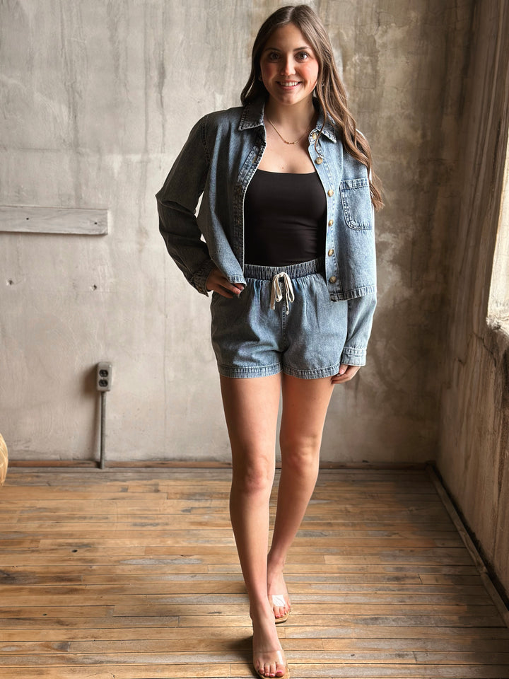 Woman wearing a denim jacket and shorts in a room with wooden floor and concrete wall.