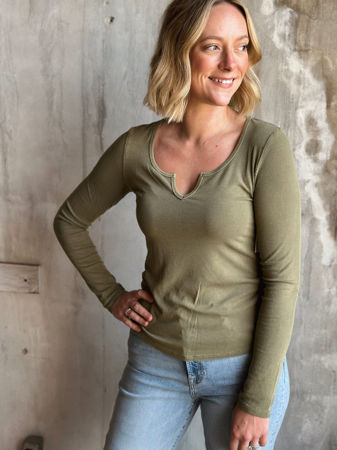 Woman wearing a green long-sleeve top and light blue jeans against a textured wall.