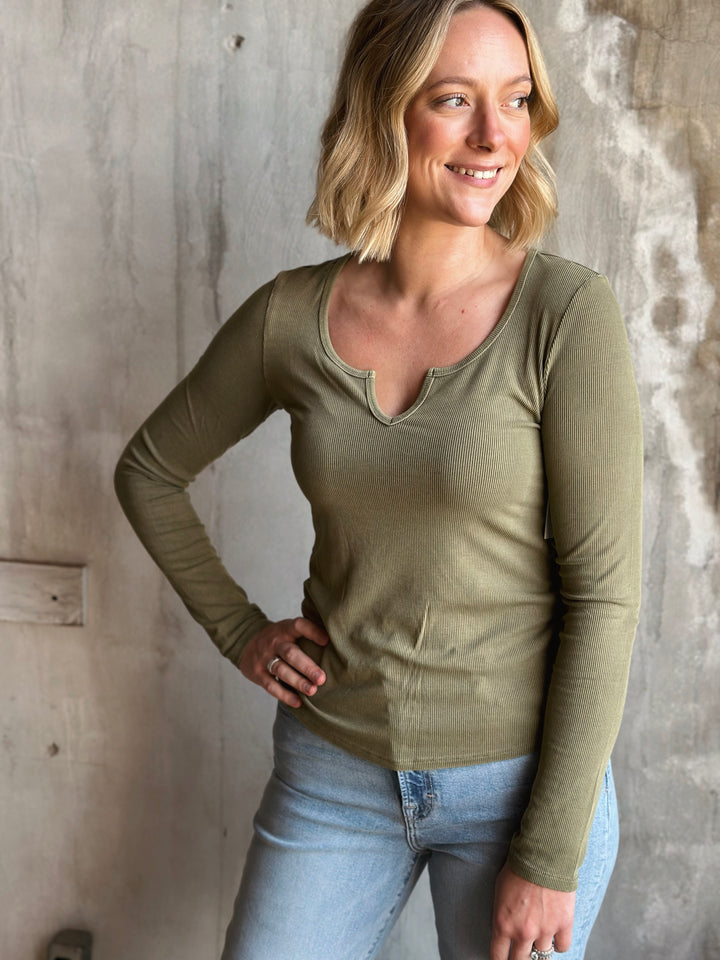 Woman wearing a green long-sleeve top and light blue jeans against a textured wall.