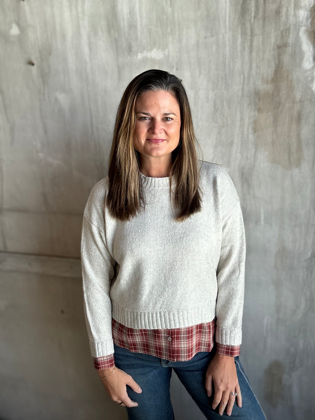 A woman with shoulder-length brown hair stands against a textured concrete wall, wearing a cream knit crewneck sweater over a red and white plaid shirt, paired with blue jeans.