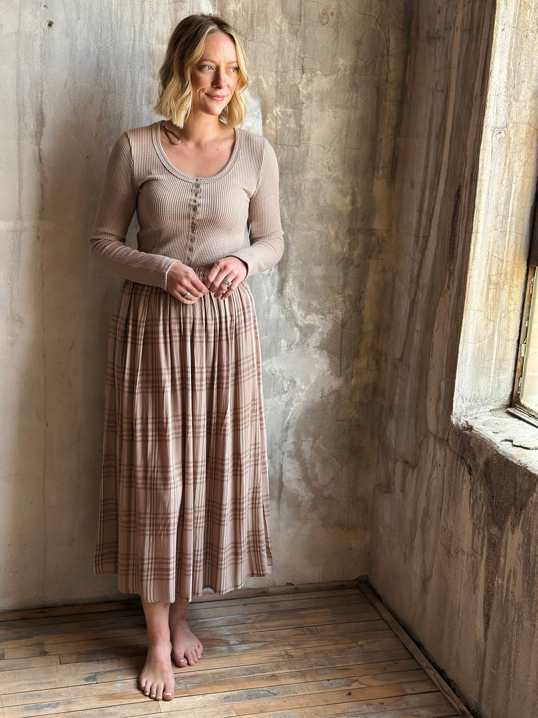 Woman wearing a beige top and plaid skirt standing against a textured wall.