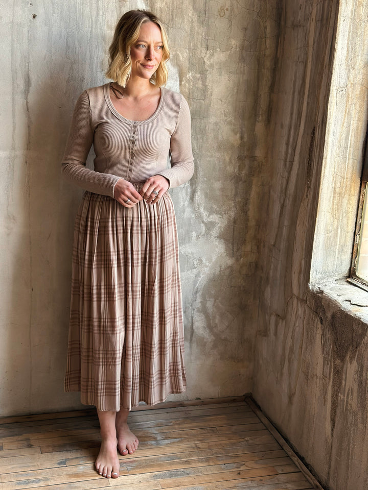 Woman wearing a beige top and plaid skirt standing against a textured wall.