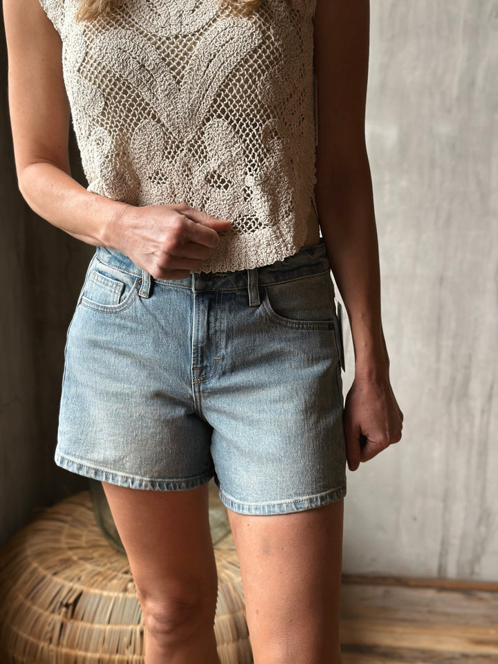 A person wearing a patterned top and light washed denim shorts against a neutral background.