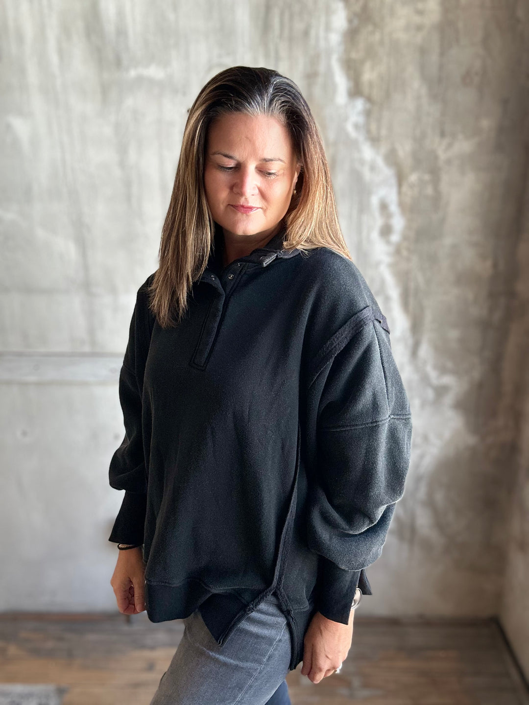 A woman wearing a dark charcoal gray oversized quarter-zip pullover sweatshirt and light gray jeans, standing against a textured concrete wall.