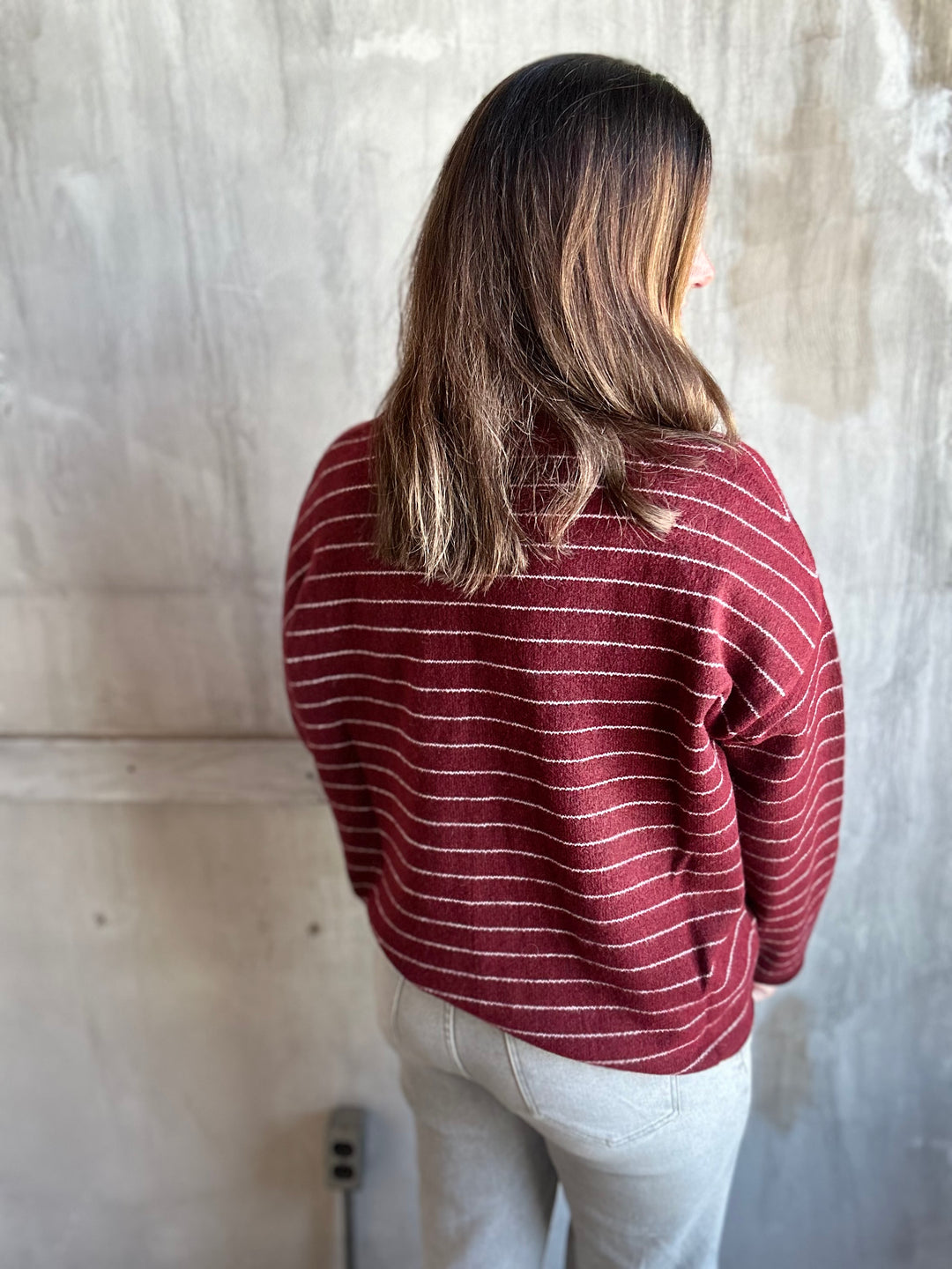 A woman viewed from behind wearing a maroon red and white striped knit sweater and light gray jeans, standing against a textured concrete wall.