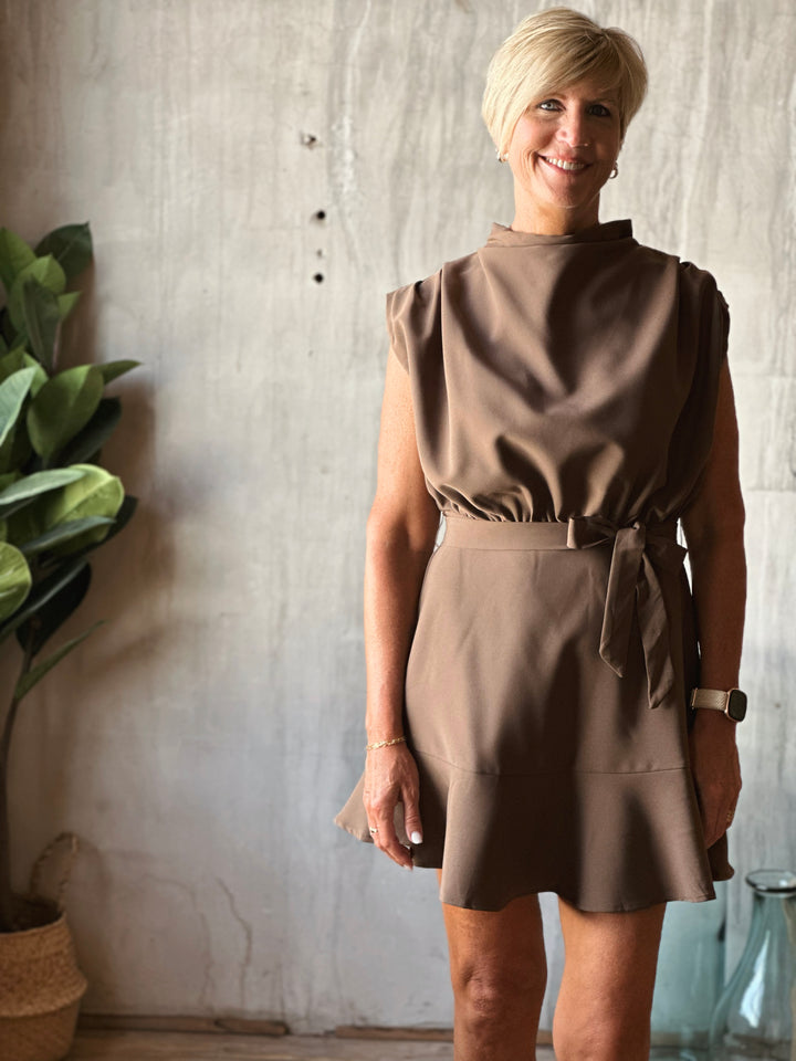 Woman wearing a brown dress standing against a textured wall with a plant to the left.