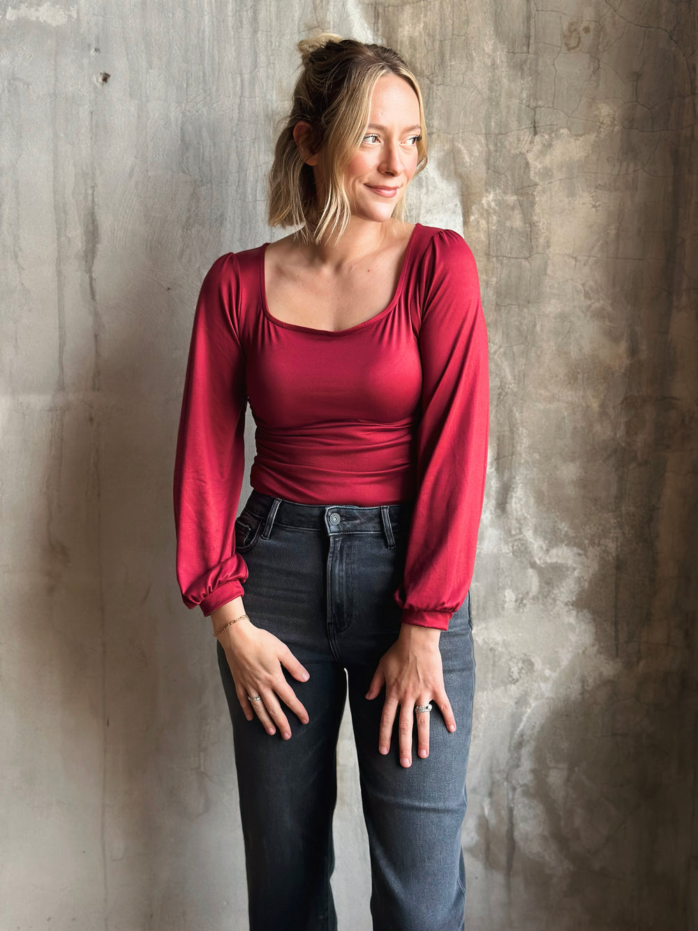 Woman wearing a red long-sleeve top and dark jeans against a textured wall.