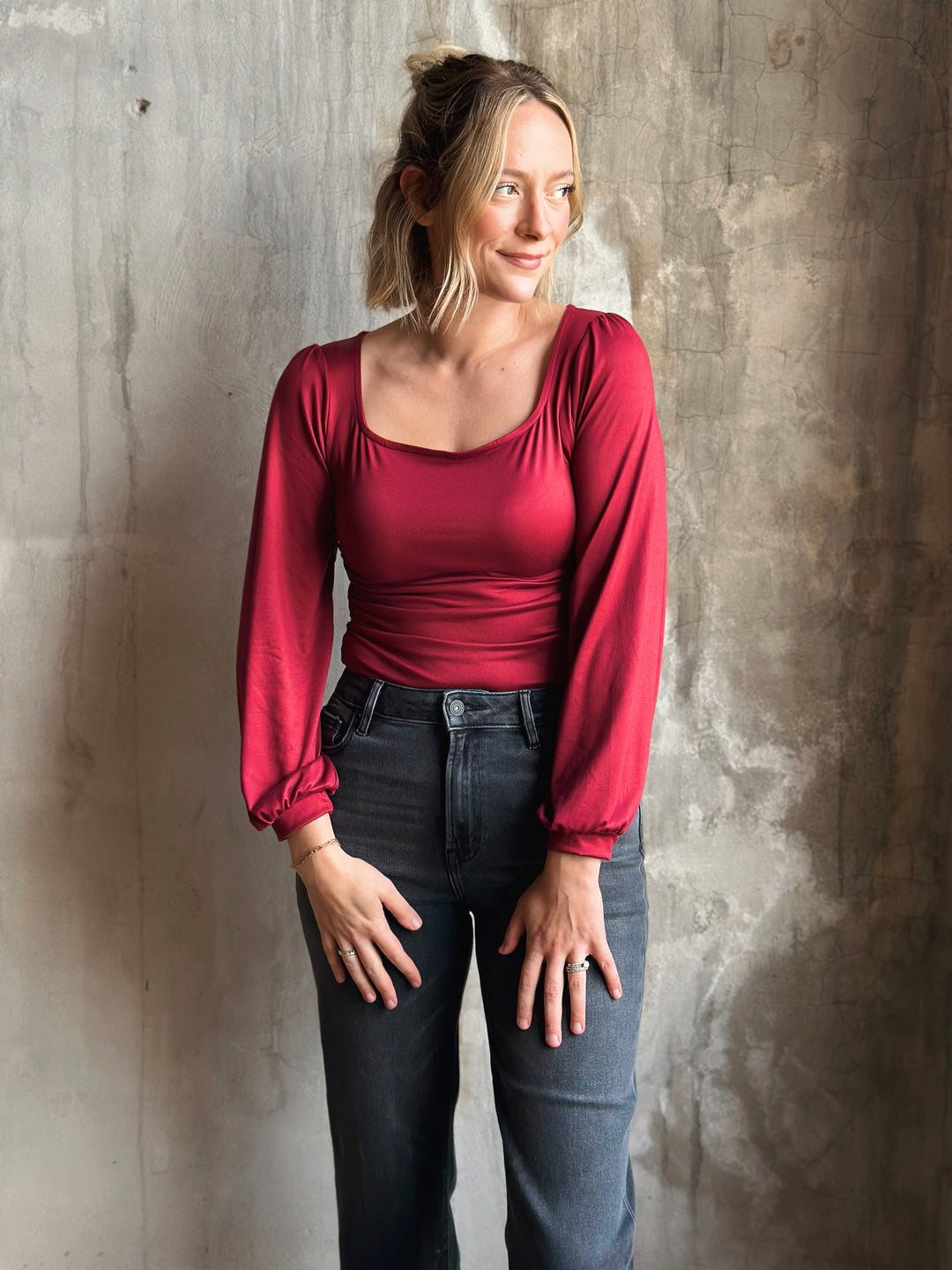 Woman wearing a red long-sleeve top and dark jeans against a textured wall.