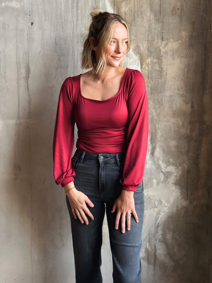 Woman wearing a red long-sleeve top and dark jeans against a textured wall.