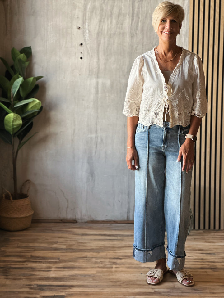 Woman wearing a white blouse and blue jeans standing indoors with a plant and wooden floor in the background.