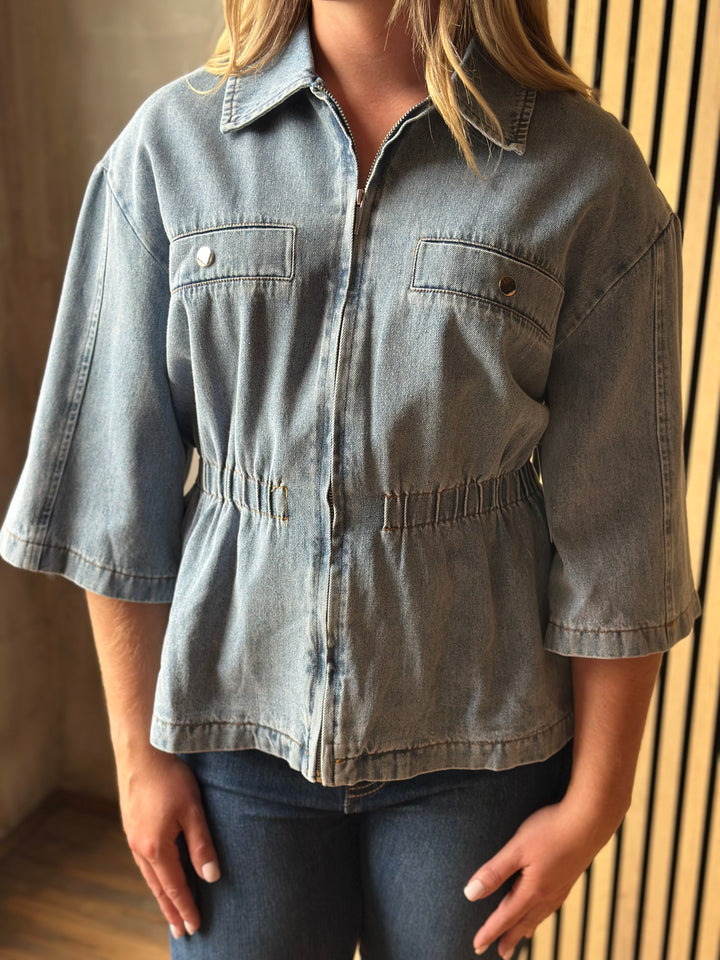 A woman wearing a light-wash blue denim shirt jacket with three-quarter sleeves, chest flap pockets, and a gathered elastic waist, paired with dark blue jeans.