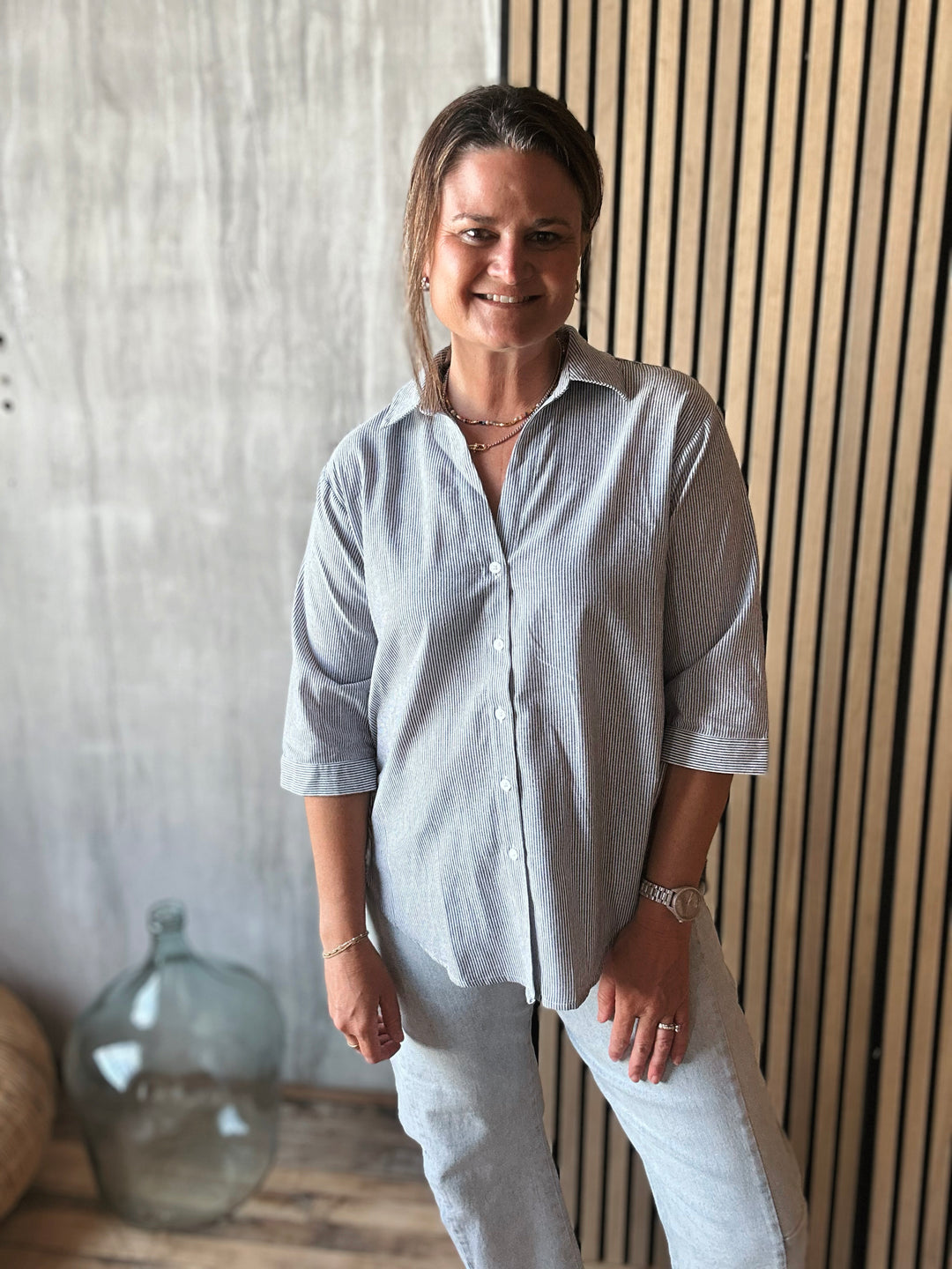 Woman wearing a light gray stripe shirt and white pants standing against a wooden wall.
