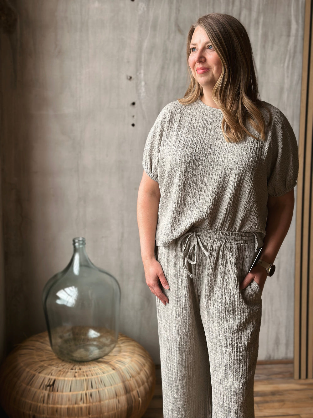 Woman wearing a sage textured outfit standing next to a large glass jar against a concrete wall.