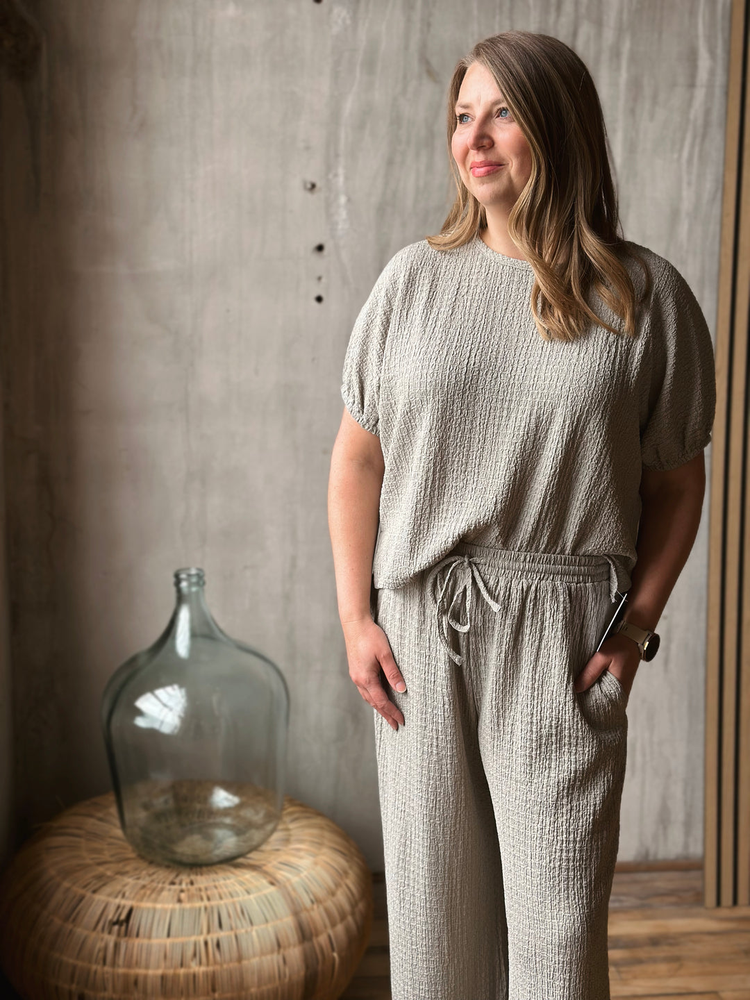 Woman wearing a sage textured outfit standing next to a large glass jar against a concrete wall.