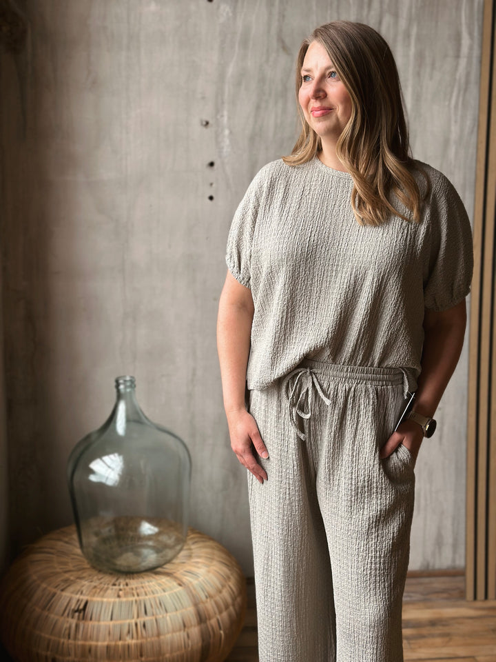 Woman wearing a sage textured outfit standing next to a large glass jar against a concrete wall.
