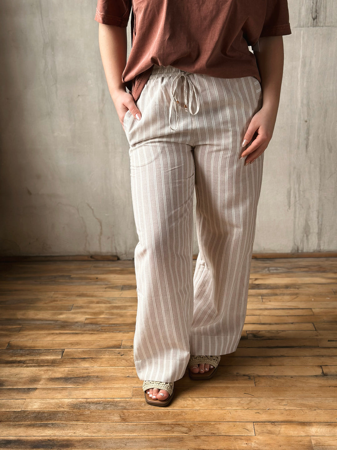 Person wearing a brown top and white striped pants standing on a wooden floor with a gray wall background.