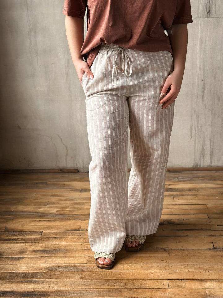 Person wearing a brown top and white striped pants standing on a wooden floor with a gray wall background.