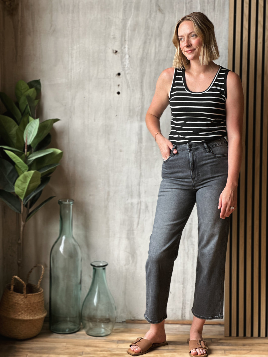 Woman wearing a black and white striped tank top and gray jeans standing in a room with decorative vases and plants.