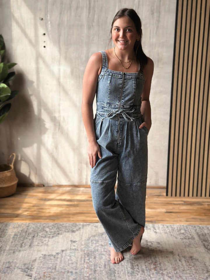 Woman wearing a denim jumpsuit standing indoors with a neutral background